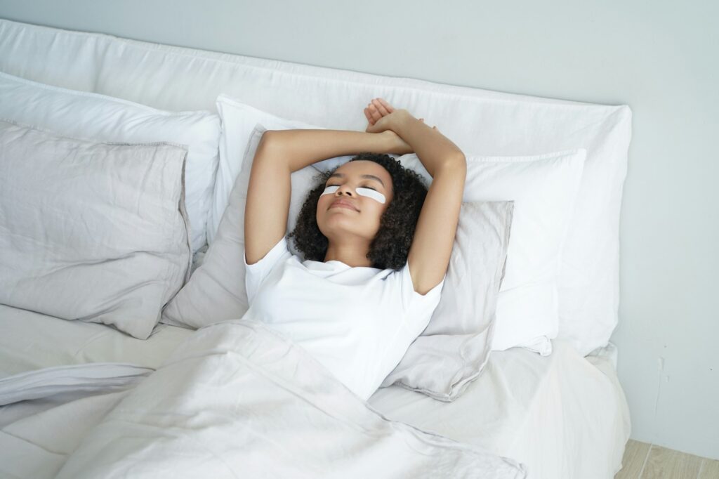 Happy afro girl sleeps with eye patches. Young woman relaxing at home in weekend morning in bed.